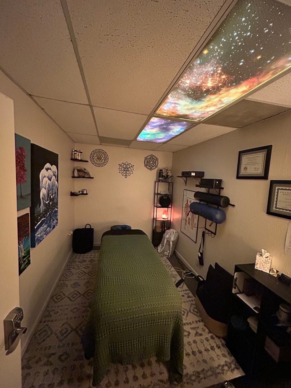 The Kinetic Freedom treatment room in Chico: massage table with a green blanket, sacred-geometry wall art, salt lamp and a lit galaxy ceiling panel